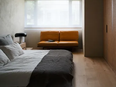 A minimalist bedroom featuring a bed with grey and white bedding, an orange lounge sofa, and wooden flooring, with a window l
