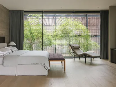 Modern, minimalist bedroom with a view of greenery outside, featuring a large window with natural light and a wooden floor.