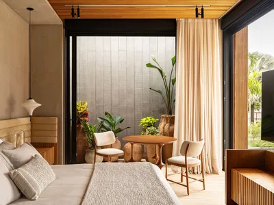 A minimalist bedroom with a bed, chairs, and a small table surrounded by potted plants, featuring hardwood floors and fabric