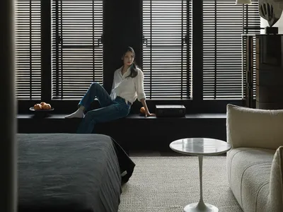 A woman sitting on a window sill in a minimalist bedroom with dark wooden window frames.