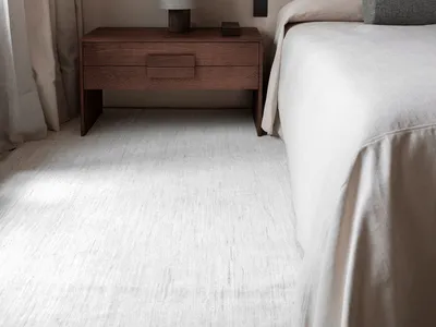 A modern minimalist bedroom featuring a bed with neutral bedding and a wooden nightstand with a lamp.