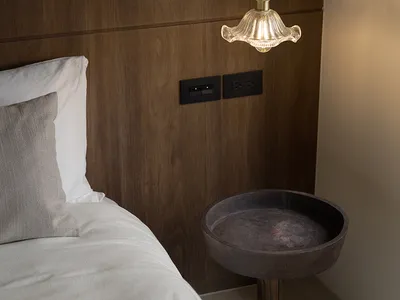 A minimalist bedroom featuring a hanging glass pendant light over a wooden bedside table, with a wooden headboard and white b