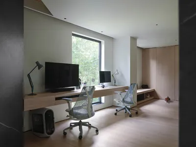 A modern home office featuring a wooden desk, ergonomic chairs, and a large window with a view of greenery.