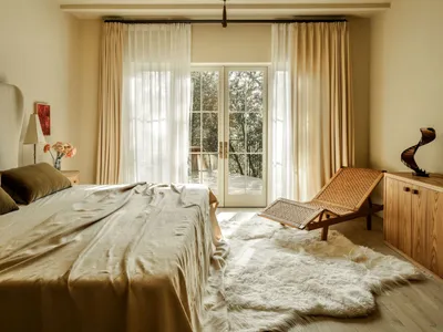 A minimalist bedroom with a bed covered in silk, large windows, and a rattan lounge chair.