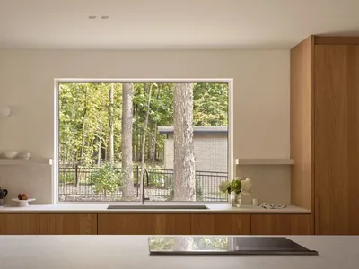A modern kitchen with wooden cabinets, a sink, and a view through a large window showing a forested area with tall trees.