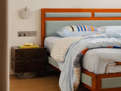 A minimalist bedroom featuring a wooden bedside cabinet and a bed with a light blue and white striped duvet, against a plain