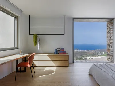 A minimalist bedroom with a wooden floor, white walls, and a large window overlooking the ocean.