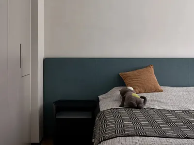 A minimalist bedroom featuring a fabric-covered bed with a wooden headboard, a small black side table, and a brown plush toy