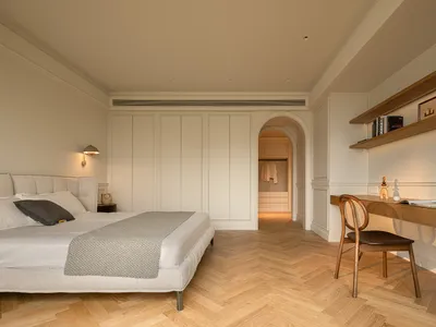 A minimalist bedroom featuring a large bed with light-colored bedding, a wooden desk with books and a plant, and a walk-in cl