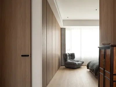 Minimalist bedroom featuring natural wood flooring and furniture, with a comfortable seating area and a window with sheer cur