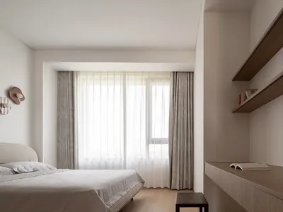 A modern bedroom with fabric headboard, wooden sides of the bed and floating desk on the wall, featuring sheer curtains over