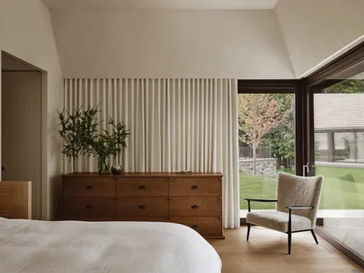 A minimalist bedroom featuring white curtains, wooden furniture, and a comfortable chair with a view of the garden.