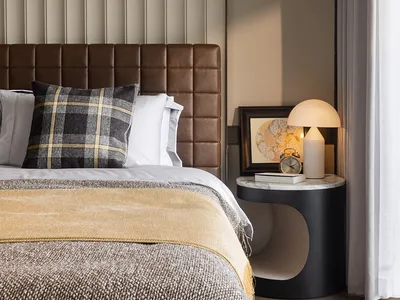 A modern bedroom with a luxurious leather headboard and checkered pillow, featuring a side table with a lamp and framed artwo