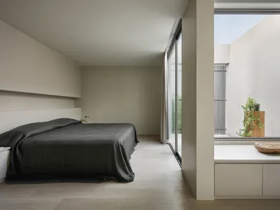 A minimalist bedroom featuring concrete flooring and walls, with a large bed covered in dark sheets and a nightstand with a g