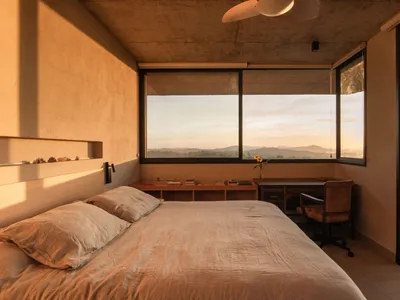 A modern minimalist bedroom with concrete walls, large windows, and a view of the mountains at sunset.