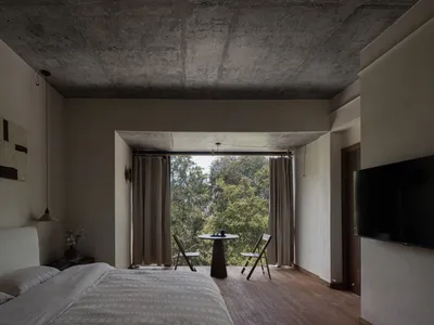 A minimalist bedroom with concrete ceiling, wooden flooring, and views of greenery outside through large sliding glass doors.