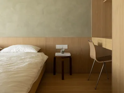 A minimalist bedroom featuring a bed with white bedding, a wooden bedside table, and a chair with a wooden backrest.