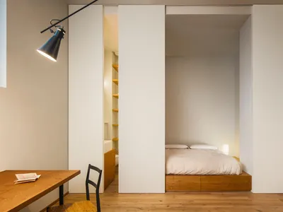 A minimalist bedroom featuring a wooden platform bed with white bedding, surrounded by white textured panels and minimalist l