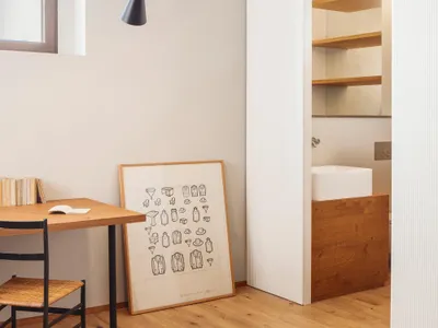 A modern home office with a wooden desk and chair, featuring a hanging lamp and a framed illustration.