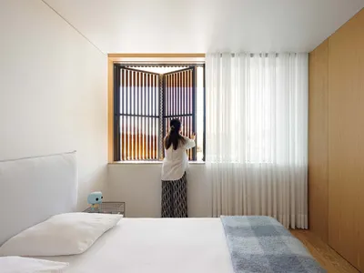 A person standing in front of a window with wooden shutters and blinds in a modern minimalist bedroom setting.