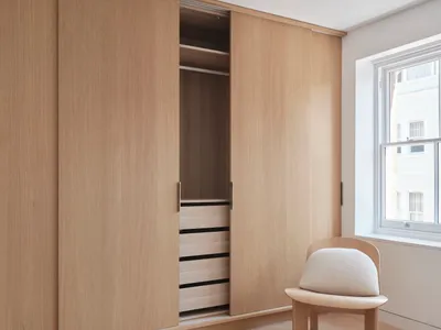 A minimalist bedroom featuring wooden cabinets with sliding doors and a white cushioned chair.