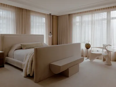 A minimalist bedroom featuring neutral-toned bedding and drapery in front of large windows with sheer curtains.