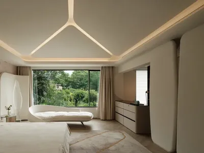 A modern bedroom with neutral colors, including a bed with a curved headboard, a white lounge chair, and a large window with