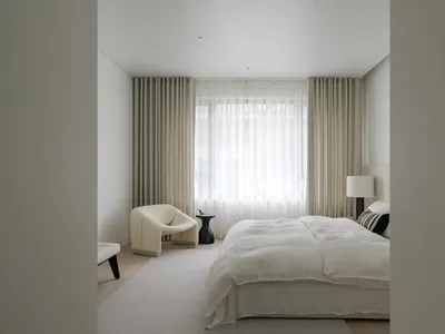 A minimalist bedroom featuring neutral-colored furniture and curtains, large windows with white sheer curtains, and a white b