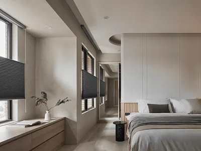 A modern minimalist bedroom featuring neutral-toned walls, a white bed with dark gray accents, and a natural light-filled win