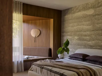 A minimalist bedroom featuring concrete and wood materials, a white bed with a striped blanket, and a wooden bench against th