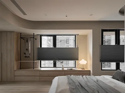 A modern minimalist bedroom with light wood flooring, a white bed with grey throw blankets, and a wooden cabinet with open sh