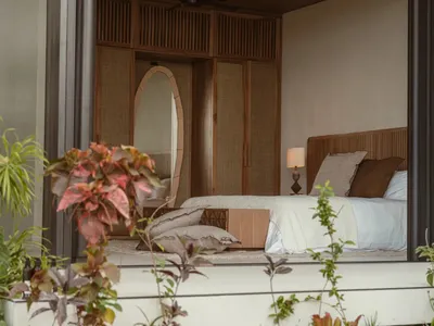 A minimalist bedroom featuring a wood frame bed with a white quilt and pillows, surrounded by green plants.