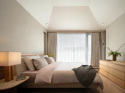 A minimalist bedroom featuring a large bed with neutral bedding, a wooden dresser, and a window with sheer curtains.