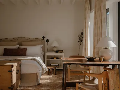 A minimalist bedroom featuring wooden furniture and neutral tones with a sunlit area and a large window.