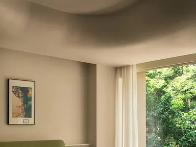 A minimalist bedroom featuring a bed with neutral fabrics, a green headboard, and a window with curtains overlooking greenery