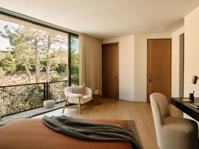 Modern minimalist bedroom featuring wood flooring, furniture, and doors with large windows overlooking nature.