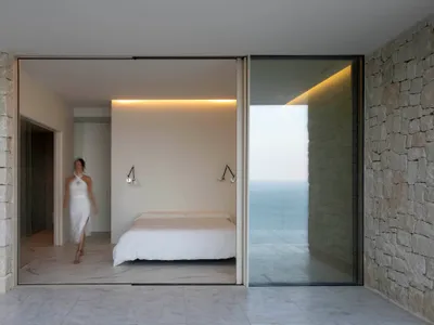 A minimalist bedroom with white walls covered in marble, featuring a single bed and a view of the ocean.