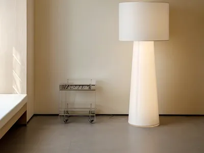 A minimalist bedroom with a large white floor lamp, a metal side table, and a bed against a beige wall.