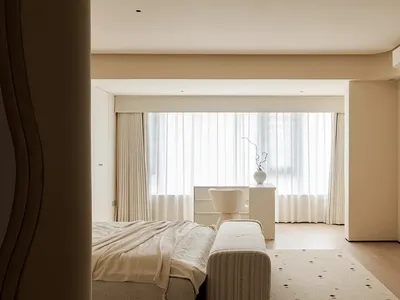 A minimalist bedroom with neutral tones and soft textiles, featuring a large bed, a small bench, and a simple desk with a vas