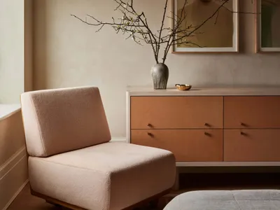 A minimalist bedroom featuring a pink armchair, a chest of drawers, and a bed with a white quilt, decorated with a vase of bl