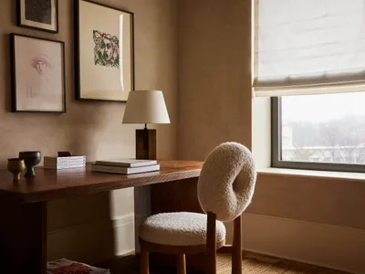 A modern home office setup featuring a wooden desk with a fabric-covered, minimalist chair, framed artworks on the wall, and