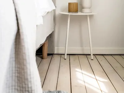A minimalist bedroom with a fabric bed against a white wall, a white side table with a vase of dried plants and a candle hold