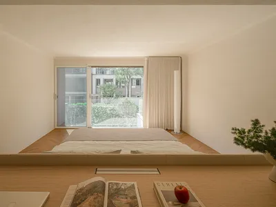 A minimalist bedroom with a low wooden frame bed, white bedding, and a window overlooking trees and buildings.