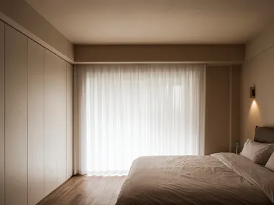 A minimalist bedroom featuring a bed with white bedding, a wall-mounted light fixture, and white curtains