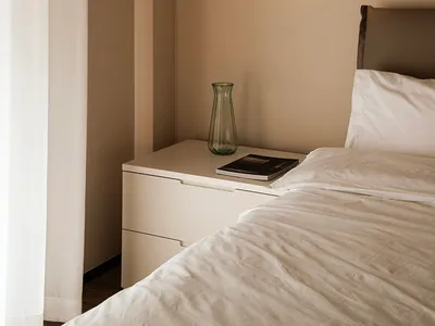 A minimalist bedroom with neutral tones featuring a bed with white bedding, a nightstand with a vase, and a wall-mounted ligh