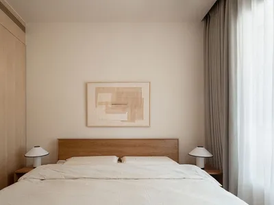 A minimalist bedroom with a wooden headboard, white bedding, and an abstract framed wall art.