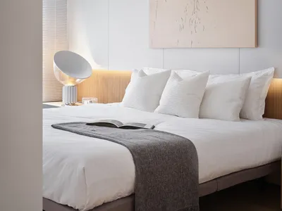 A minimalist bedroom with white bedding, a grey throw blanket, and a peach-colored abstract painting hanging above the bed. T