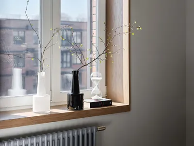 A minimalist bedroom with a wooden window sill decorated with vases and branches, featuring a radiator and a carpet on the fl