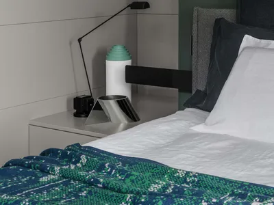 A minimalist bedroom with concrete, metal, and fabric materials, featuring a bed with a green patterned blanket, a modern lam