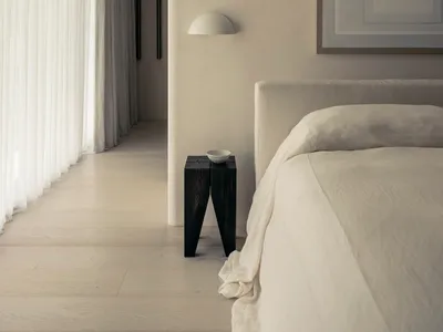 A minimalist bedroom featuring a white fabric upholstered bed, a wooden nightstand, and light gray curtains on a large window
